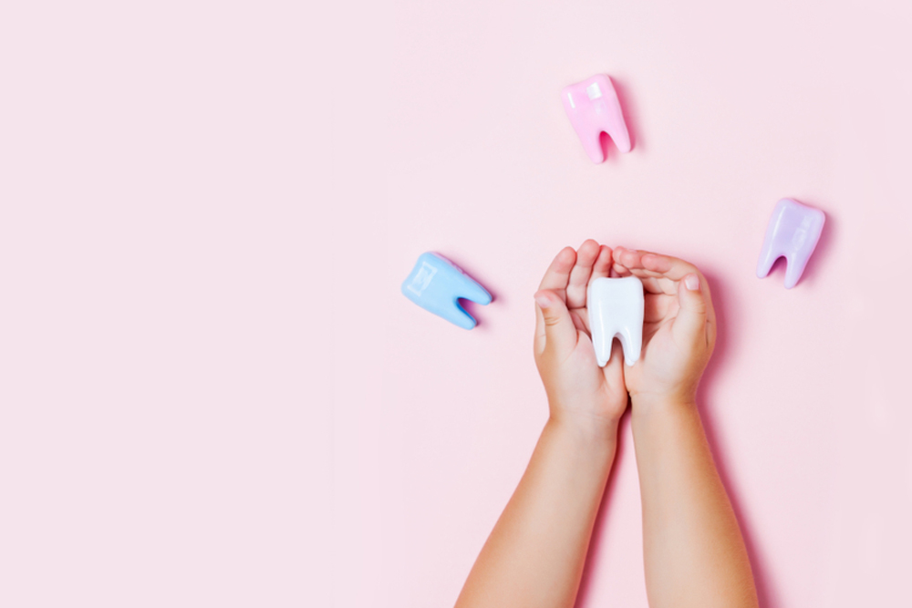 Pink background. Child's hands holding a white tooth-shaped floss container with three other tooth-shaped floss containers around the hands.